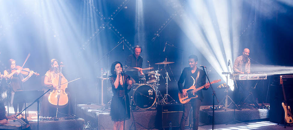 This live concert photo shows a glamorous Ninet in black dress singing into a mic, with Dudu in black business-casual garb playing guitar next to her